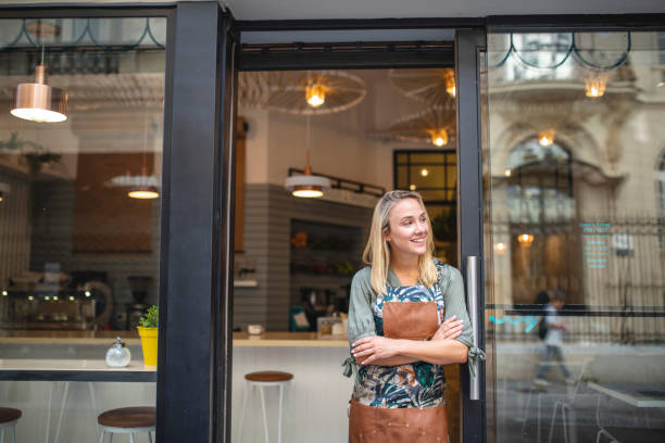 Hopeful Argentine female cafe owner standing in apron with arms crossed at open front door and looking away from camera.